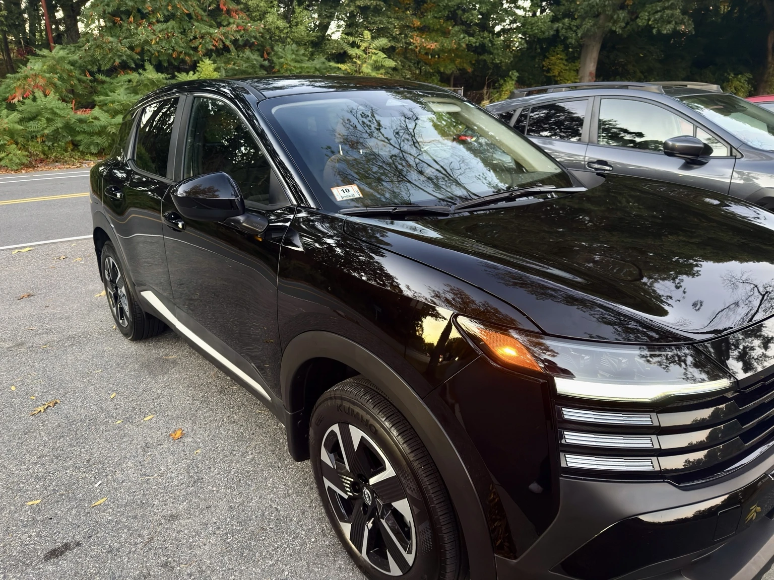 Owner's black 2025 Nissan Kicks SV AWD viewed from the front three-quarter angle on a tree-lined road beside another parked car